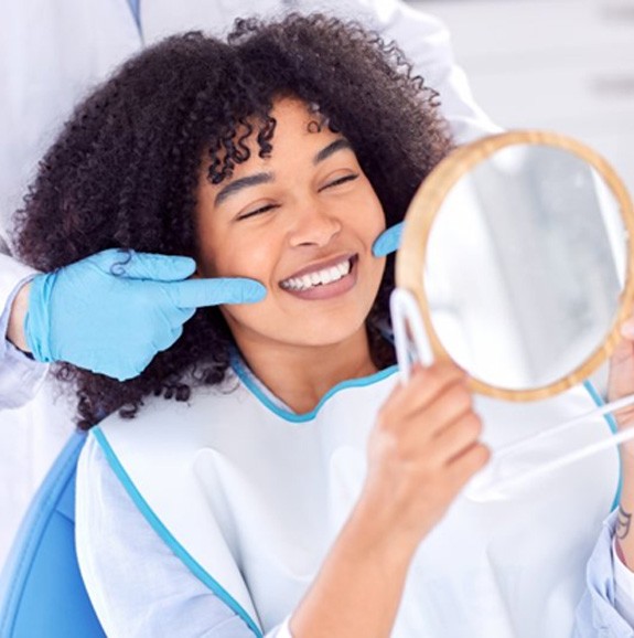 Dentist pointing at patient’s smile as she examines her teeth in small mirror